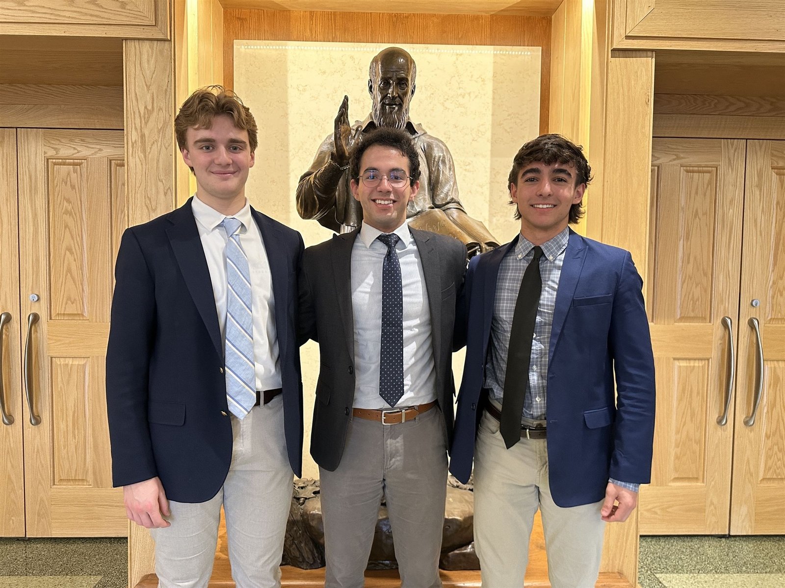Three Salesianum students in blazers standing with a bronze statue in a wood-paneled hall